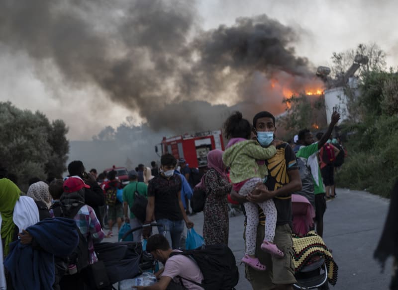 Prchající rodiny migrantů, za zády jim hoří jejich původní útočiště, uprchlický tábor Moria. 