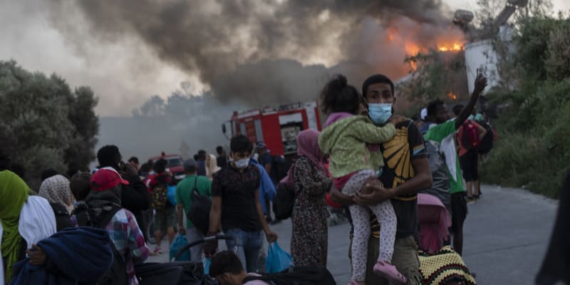 Prchající rodiny migrantů, za zády jim hoří jejich původní útočiště, uprchlický tábor Moria. 