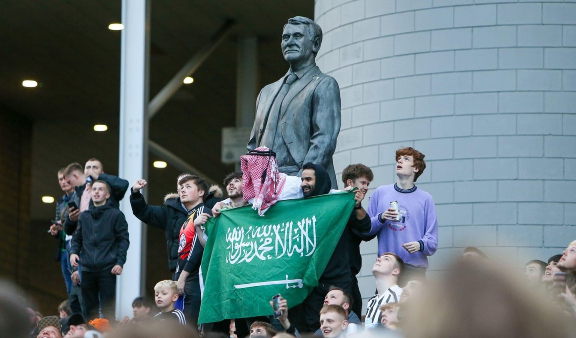 Skupina fanoušků Newcastlu se saúdskoarabskou vlajkou u sochy trenérské ikony Bobbyho Robsona. Ne všichni ale mají z nových majitelů klubu stejnou radost.