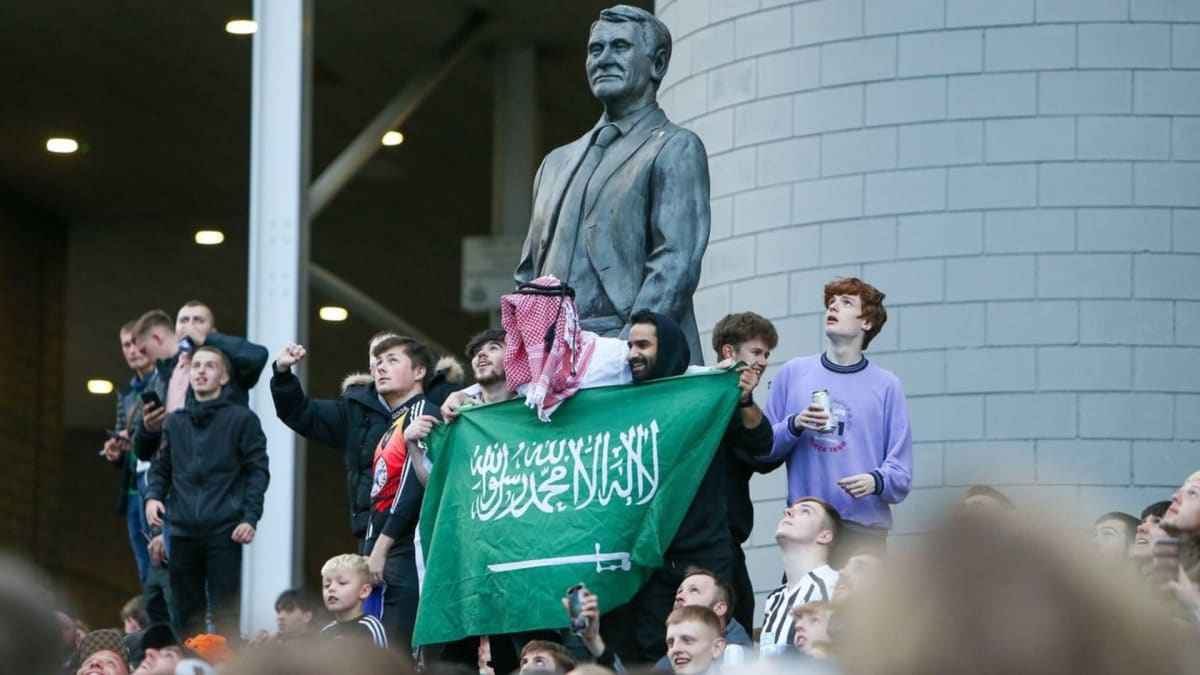 Skupina fanoušků Newcastlu se saúdskoarabskou vlajkou u sochy trenérské ikony Bobbyho Robsona. Ne všichni ale mají z nových majitelů klubu stejnou radost.