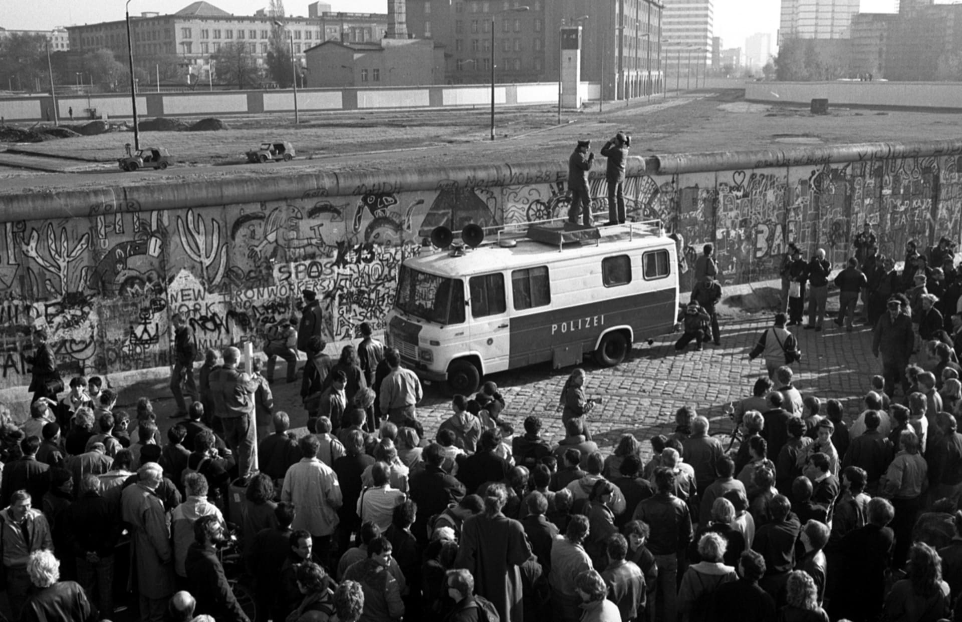 Pohled do pásma nikoho ze západoberlínské strany zdi. Uvnitř území nikoho jsou dobře vidět dva „obrněné“ trabanty východoněmeckých pohraničníků