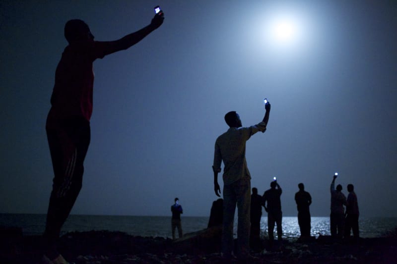 Afričtí migranti na pobřeží nočního Džibuti City zvedají telefony do výšky ve snaze zachytit poměrně levný signál ze sousedního Somálska a navázat tak alespoň nějaké spojení s příbuznými  v zahraničí.