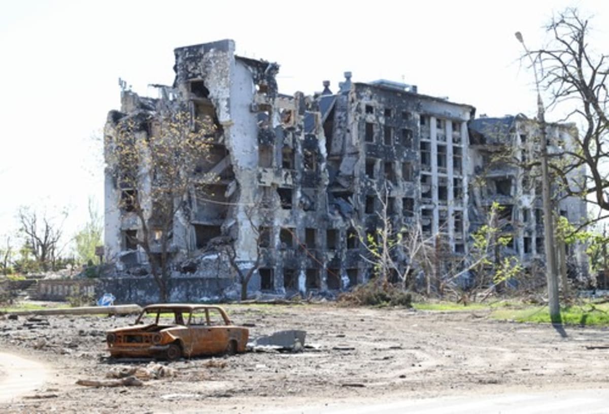 Ohořelý vrak auta před zničenými obytnými budovami v Mariupolu