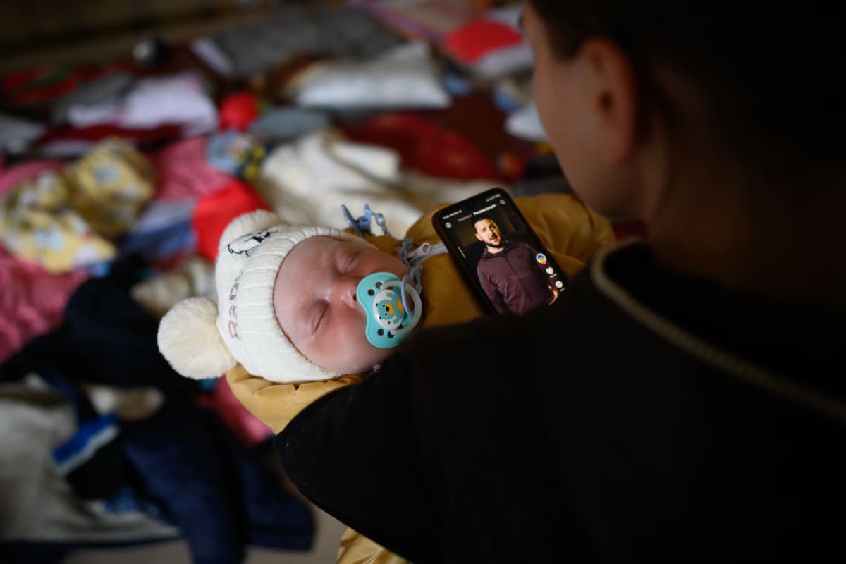 Žena sleduje projev pezidenta Volodymyra Zelenského, zatímco jí v náručí spí dcera. Snímek byl pořízen v zařízení pro uprchlíky ve Lvově