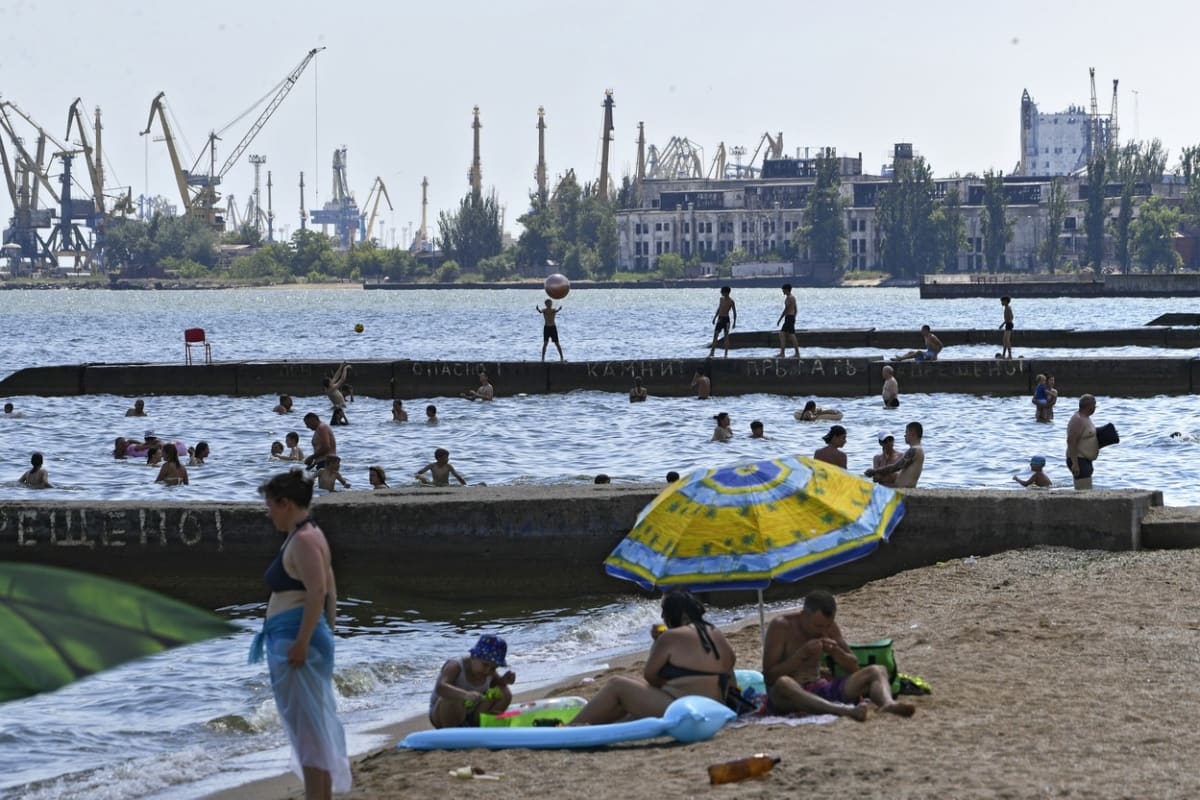 Lidé si užívají pěkného počasí na mariupolské pláži. V pozadí jsou trosky oceláren Azovstal