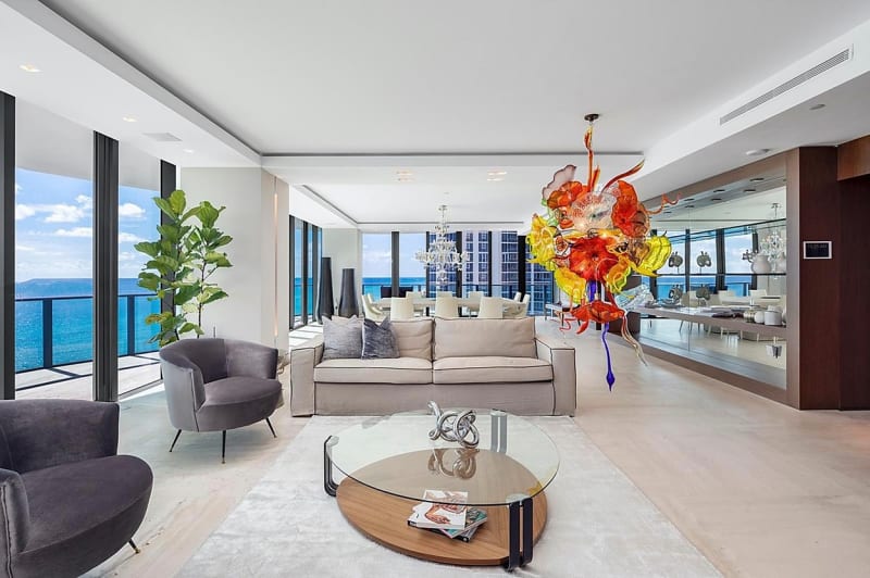 Jak bydlí Lionel Messi v Miami: V obývacím pokoji jsou velká okna sahající od podlahy až ke stropu, centrem místnosti jsou dvě světlé pohovky doplněné o stejně neutrální křesla, jako kontrast zde slouží barevná instalace na stropě.