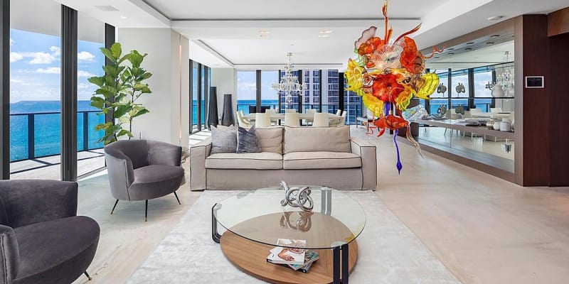 Jak bydlí Lionel Messi v Miami: V obývacím pokoji jsou velká okna sahající od podlahy až ke stropu, centrem místnosti jsou dvě světlé pohovky doplněné o stejně neutrální křesla, jako kontrast zde slouží barevná instalace na stropě.