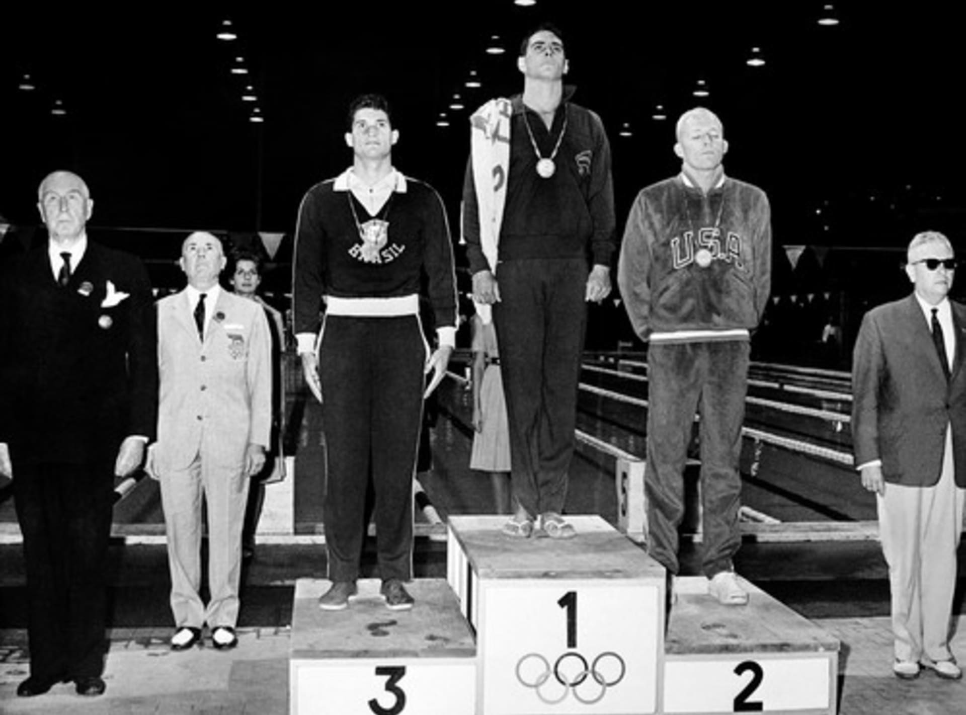 Vítěz zlaté medaile John Devitt z Austrálie, vedle něj stříbrný medailista Lance Larson (vpravo) a bronzový medailista Manuel Dos Santos poslouchají státní hymnu na pódiu během ceremoniálu udílení cen za plavání.