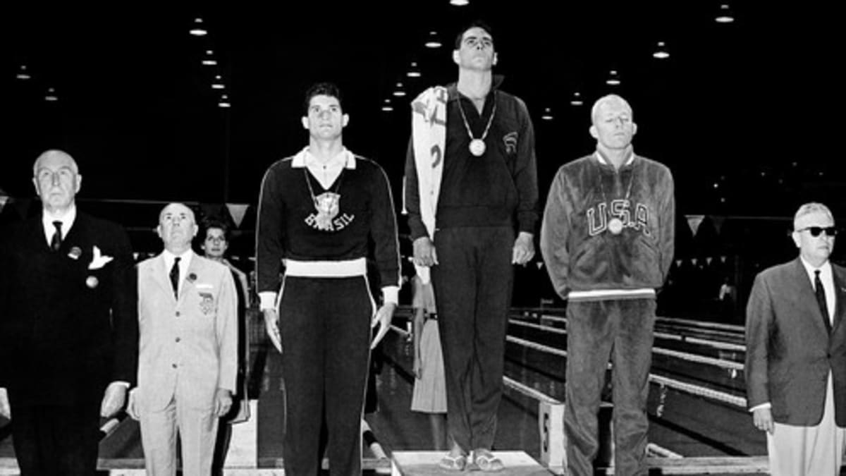 Vítěz zlaté medaile John Devitt z Austrálie, vedle něj stříbrný medailista Lance Larson (vpravo) a bronzový medailista Manuel Dos Santos poslouchají státní hymnu na pódiu během ceremoniálu udílení cen za plavání.
